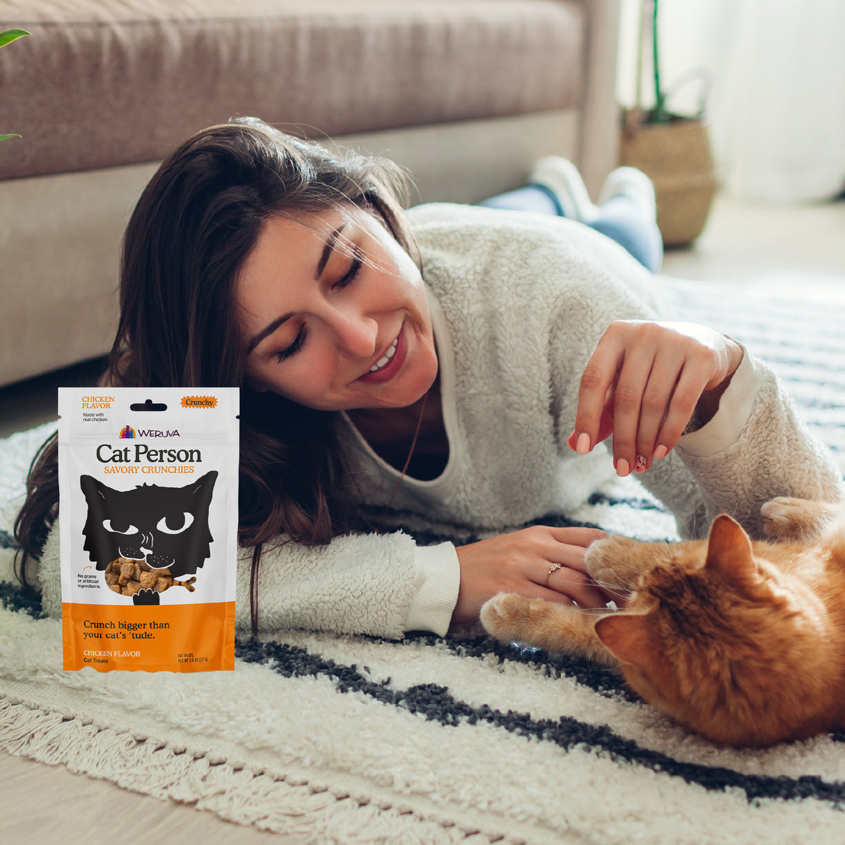 A woman lies on a rug indoors, smiling and petting an orange cat. In the foreground, a bag of Cat Person Chicken Savory Crunchies is visible next to her hand.