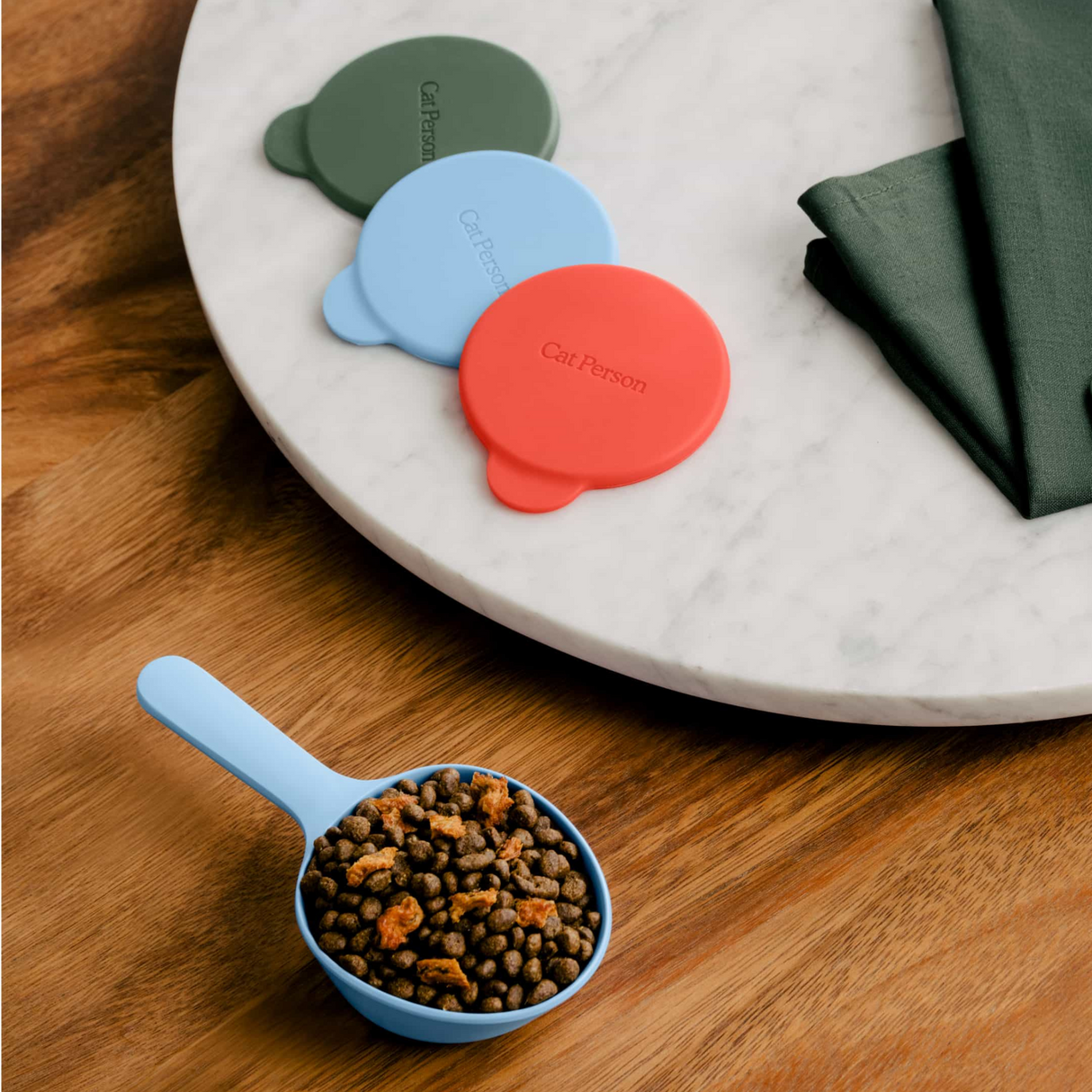 The Cat Person Serve & Store Set by Cat Person features a blue portion control scoop with dry cat food, placed on a wooden surface beside three airtight lids in green, blue, and red, plus a dark green napkin on a round marble tray.