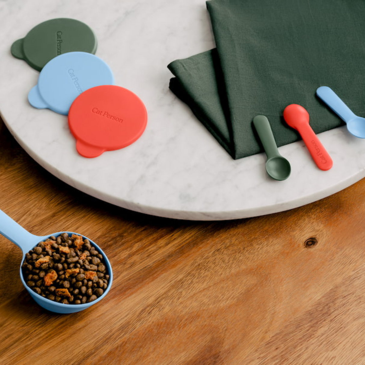 The Cat Person Serve & Store Set by Cat Person features a blue portion control scoop with dry cat food, displayed on a table beside green napkins, two small spoons, and three colorful airtight lids—perfect silicone accessories for cats.