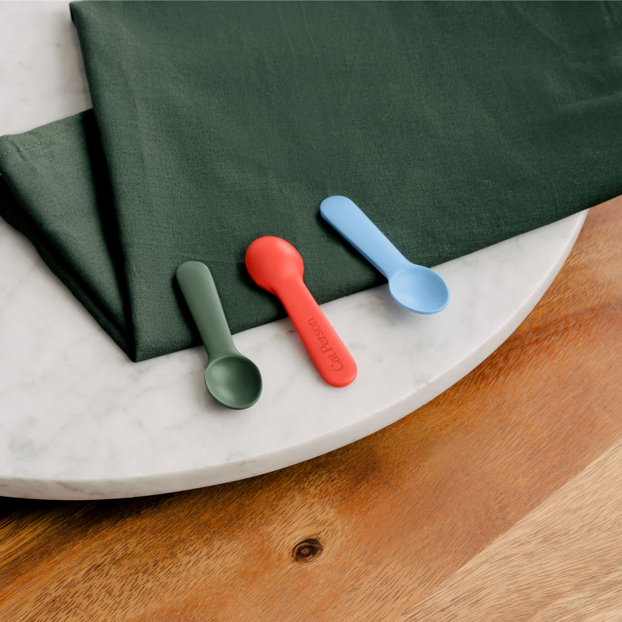The Cat Person Serve & Store Set by Cat Person, featuring three small silicone scoops in green, red, and blue, is arranged side by side on a green napkin atop a marble table with part of a wooden surface in view.