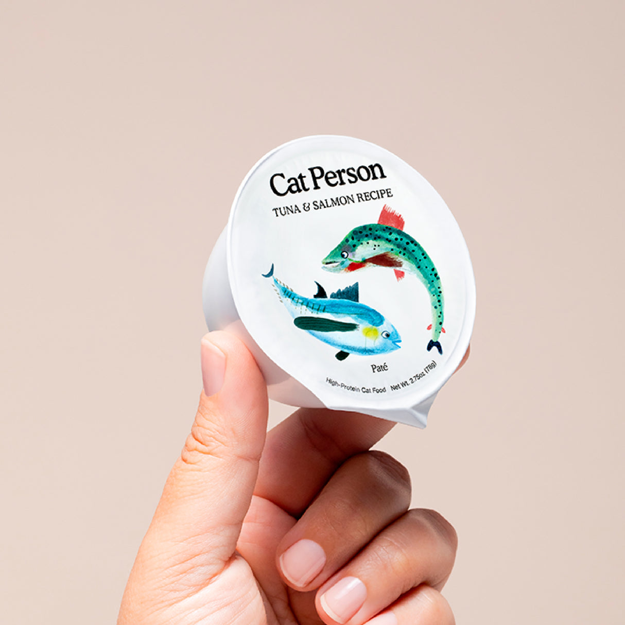 A hand holds a small container of Cat Person Tuna & Salmon Paté, a protein-rich cat food, against a beige background. The label shows charming blue tuna and green salmon illustrations.