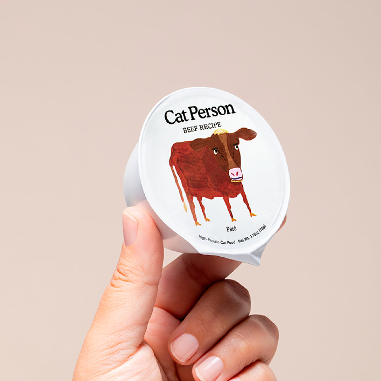 A hand holds a small container of Cat Person Beef Shreds in Broth cat food with a brown cow illustration on the lid, set against a plain beige background, showcasing high-quality beef for balanced nutrition.