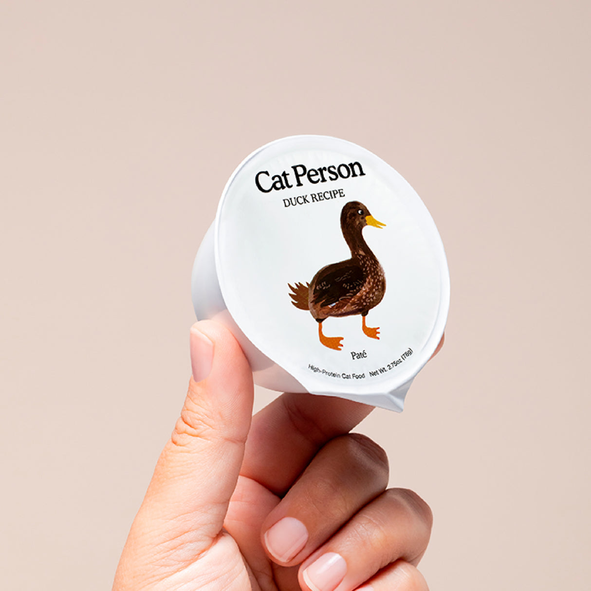 A hand holds a small container of Cat Person Duck Shreds in Broth cat food, with a duck illustration on the lid set against a plain, light background.