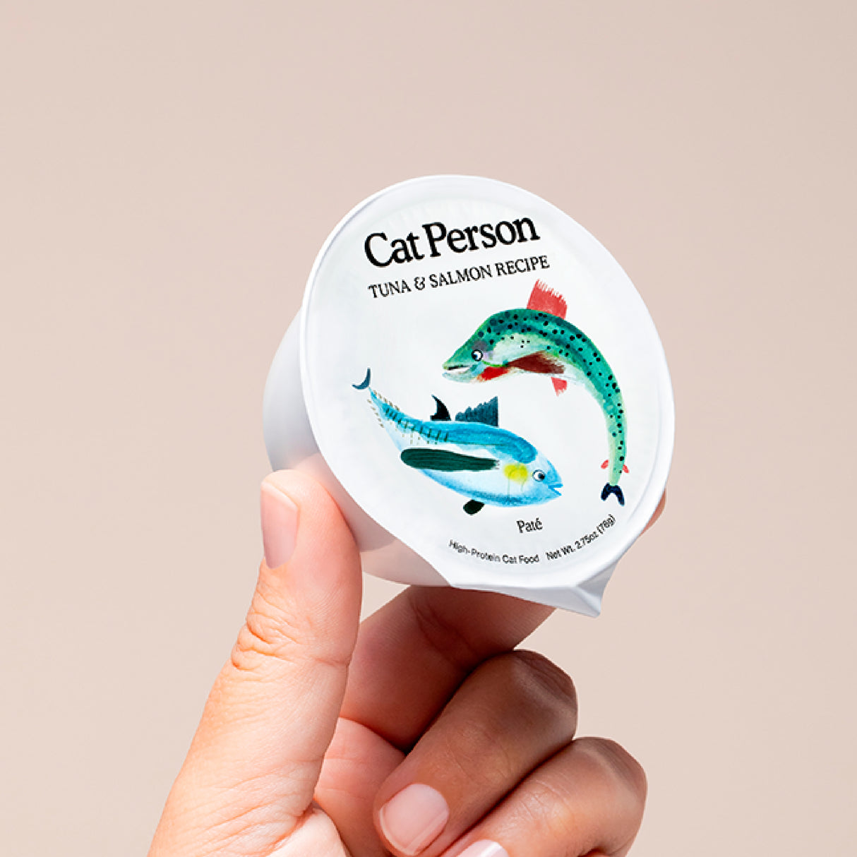 A hand holds a small container labeled Cat Person Salmon & Mackerel Paté, featuring illustrations of salmon and mackerel on the lid. This cat food offers protein-rich, balanced nutrition for your feline against a neutral background.