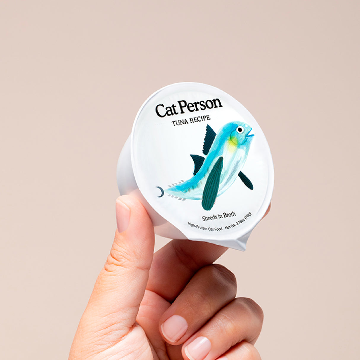 A hand holds a small container of Cat Person Tuna Shreds in Broth cat food, featuring an illustration of a blue fish on the label against a plain beige background.