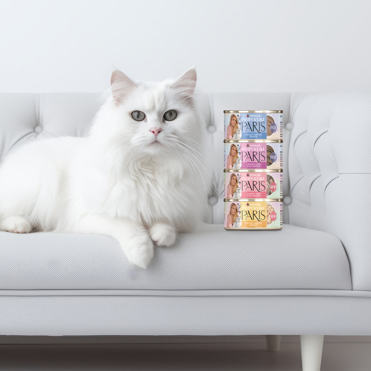 A fluffy white cat lounges on a white tufted sofa next to four Weruva Salmon and Tuna Dinner in Gravy cans, in a minimalist, bright setting that highlights the delicious gravy and wild-caught salmon flavors.