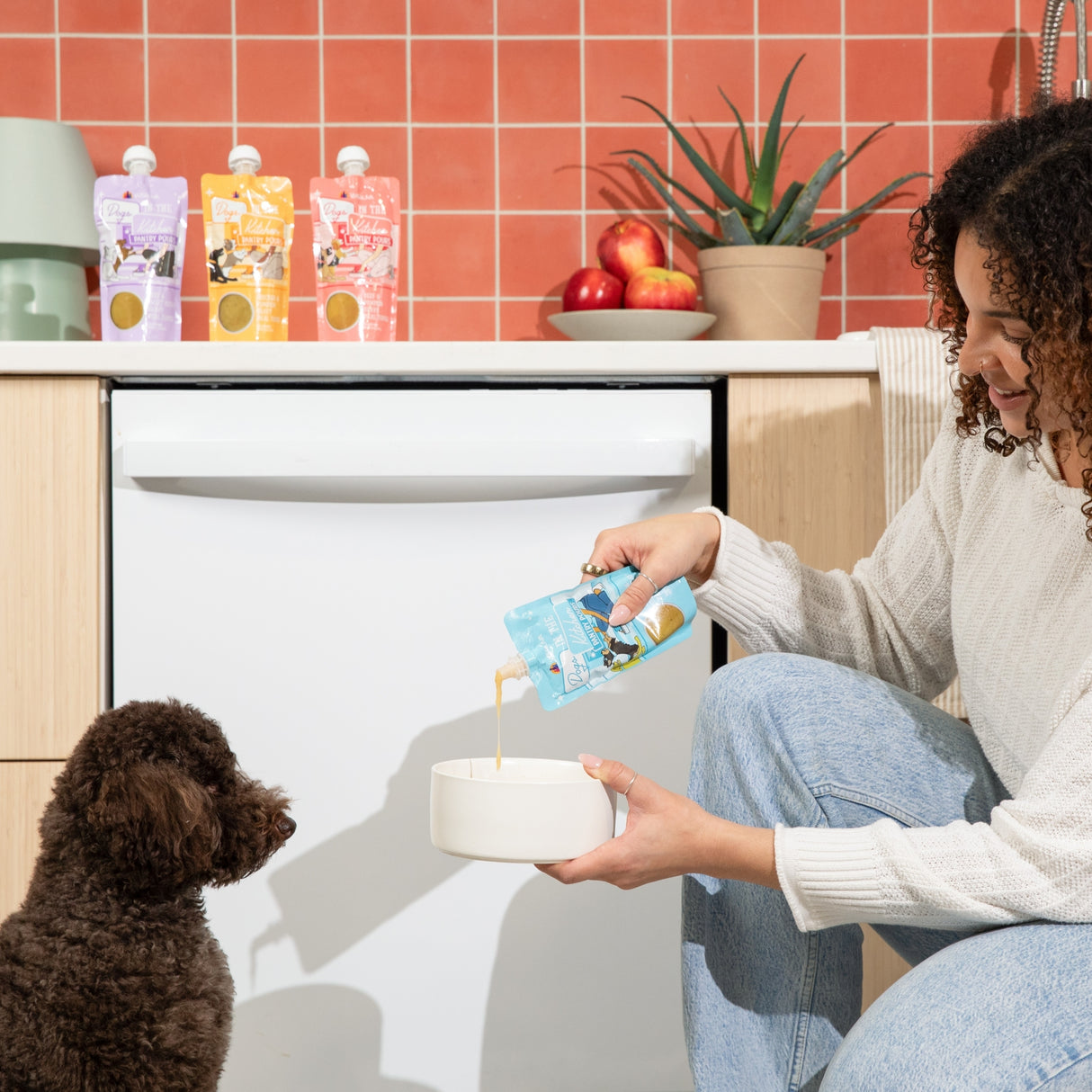 In a kitchen with red tiled walls and plants, a woman pours Dogs in the Kitchen Chicken & Sweet Potato Gravy Meal Topper from a pouch into a bowl for her small brown dog, while three pouches are visible in the background.
