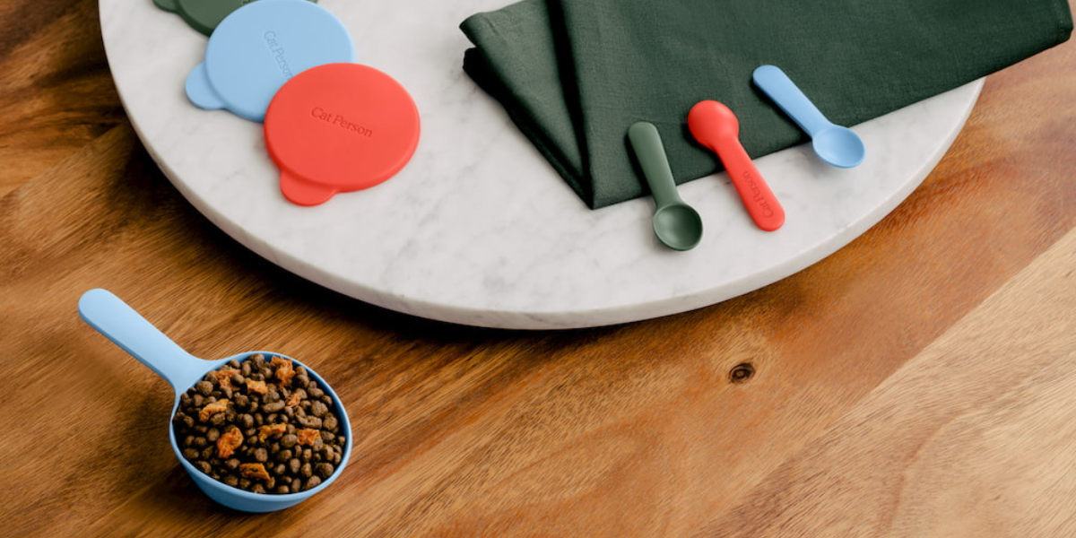 A blue scoop of dry cat food sits on a wooden table near a round marble slab with blue and red silicone can covers, green napkins, and small blue and green spoons.