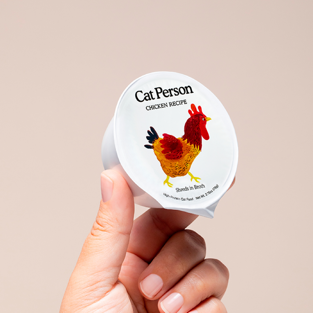 A hand holds a small cup of Cat Person Chicken Shreds in Broth, topped with a chicken illustration on the lid. This premium cat food offers balanced nutrition and is set against a simple, light background.