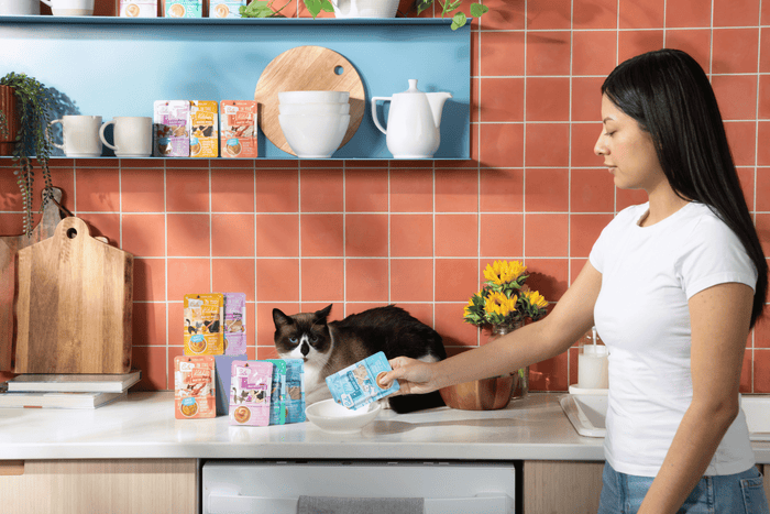 A woman in a white shirt pours food from a pouch into a bowl for a black and white cat sitting on a kitchen counter, with cat food pouches, sunflowers, and kitchenware in the background.