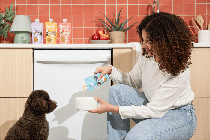 A woman kneels in a kitchen, pouring food from a pouch into a bowl for a seated brown dog, with colorful pouches and fruit on the counter behind her.