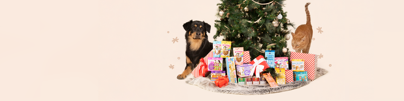 A puppy and a cat stand next to a decorated Christmas tree surrounded by assorted pet treats and wrapped gifts on a white fur rug.